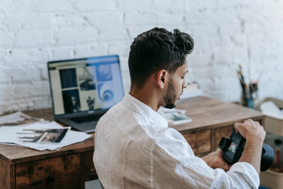 Side view focused young ethnic male photographer wearing casual clothes looking through pictures in modern photo camera while sitting near with laptop in light office