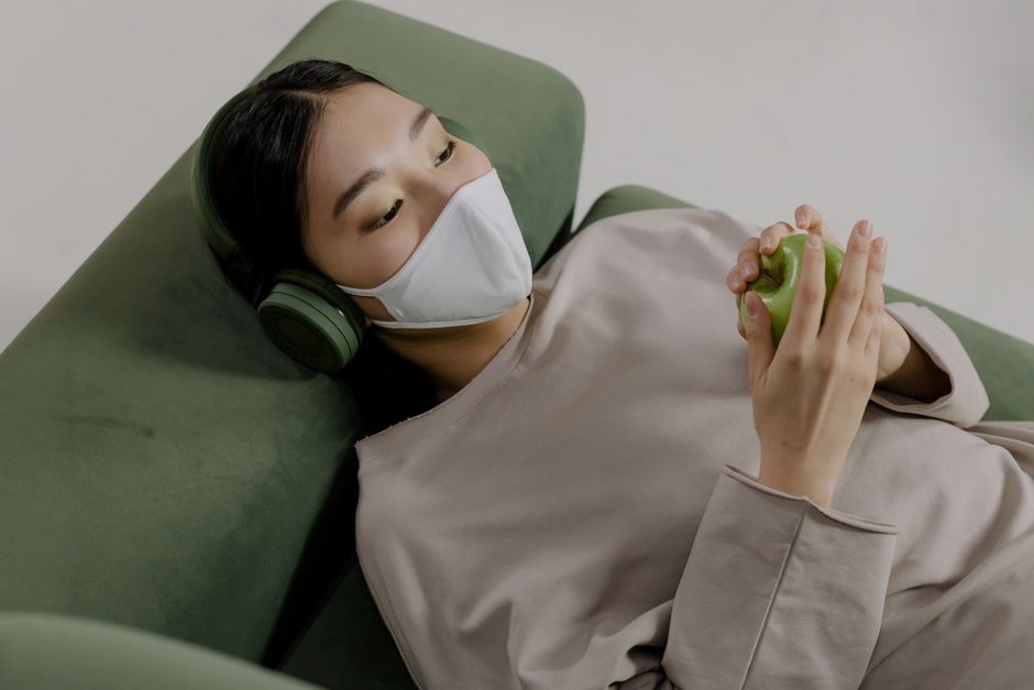Asian woman relaxing indoors with a face mask, headphones, and a green apple.