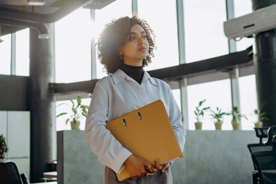 A professional woman stands in a stylish office, holding a folder with natural light streaming in.