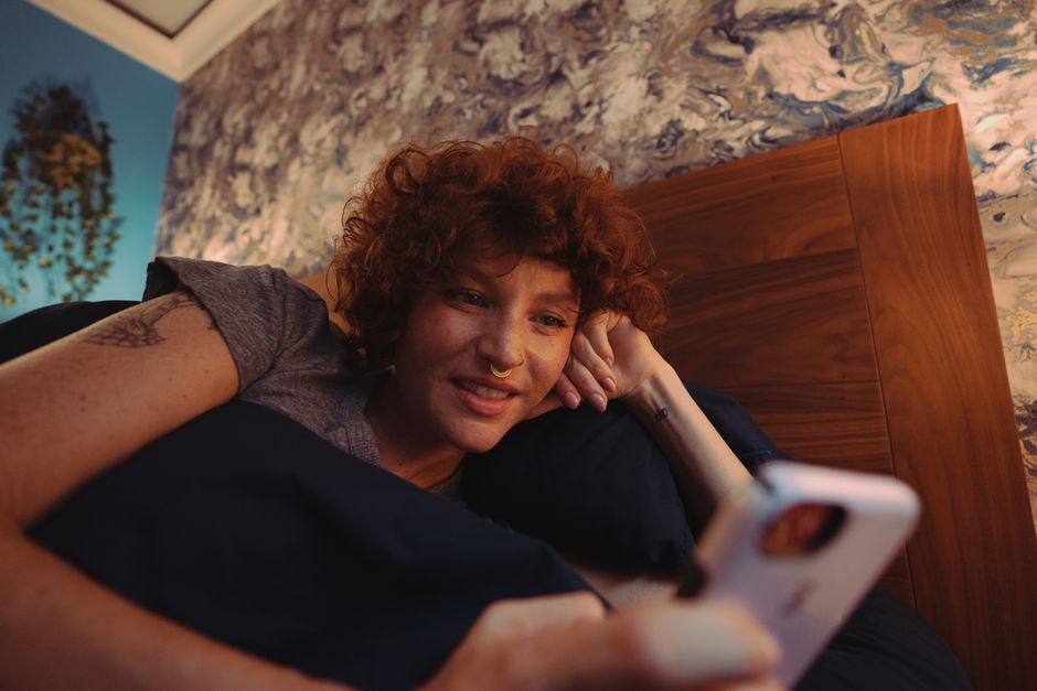 A woman with curly red hair smiles while using a smartphone in bed. Cozy indoor setting.