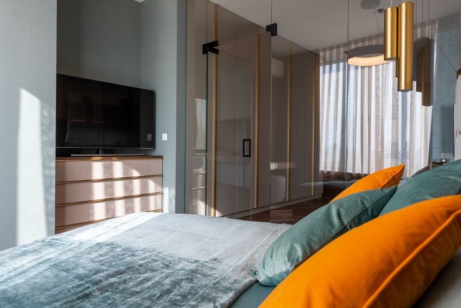 Interior of modern bedroom with colorful pillows on bed placed against TV with drawer near glass door in contemporary apartment