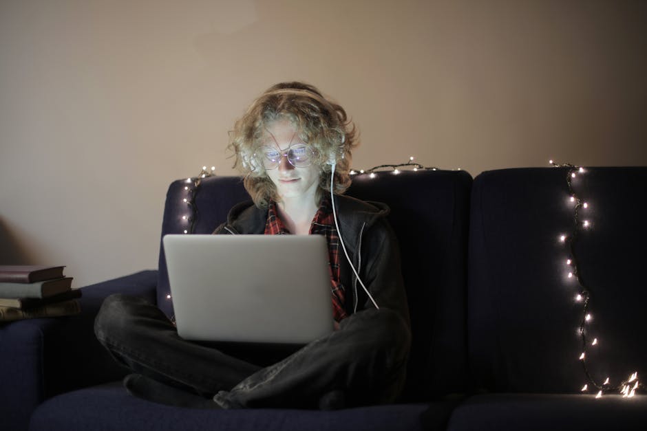 Enjoyable teen boy in casual outfit eyeglasses sitting on sofa with luminous garland browsing laptop and listening to music on headphones during evening at home